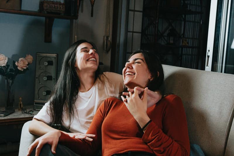 two women sit together on a couch and laugh